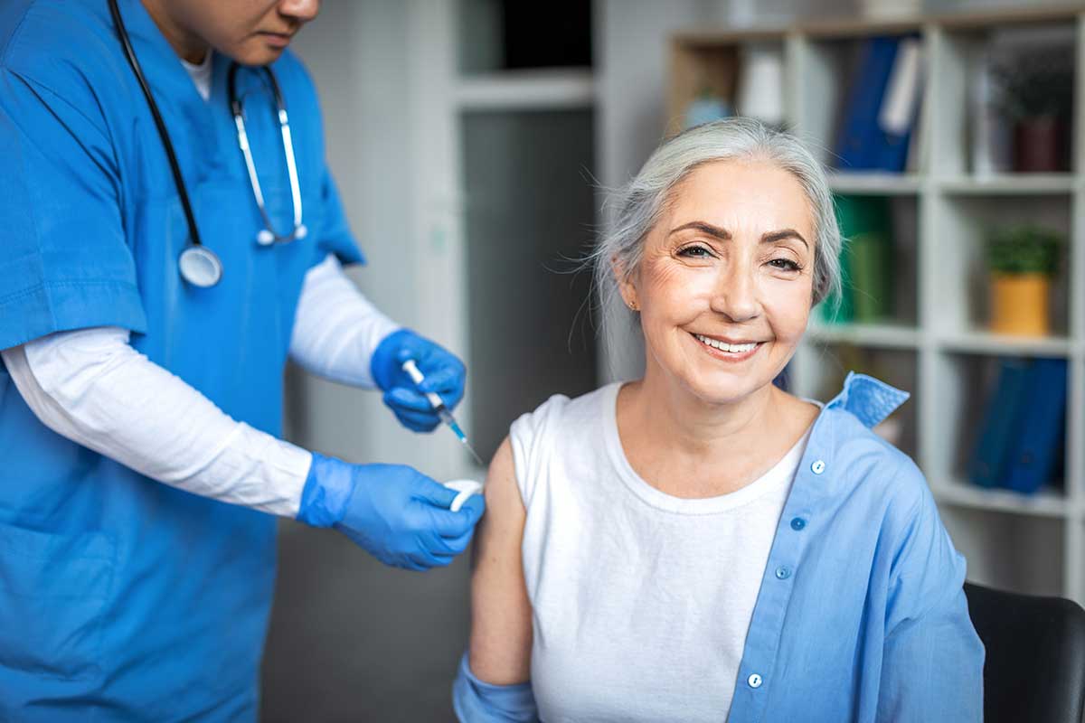 Smiling woman older woman receives vaccination from medical professional who is mostly offscreen.