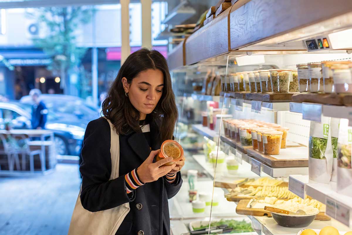 Young woman in coat stands at grocery display examining the label on a packaged food item.