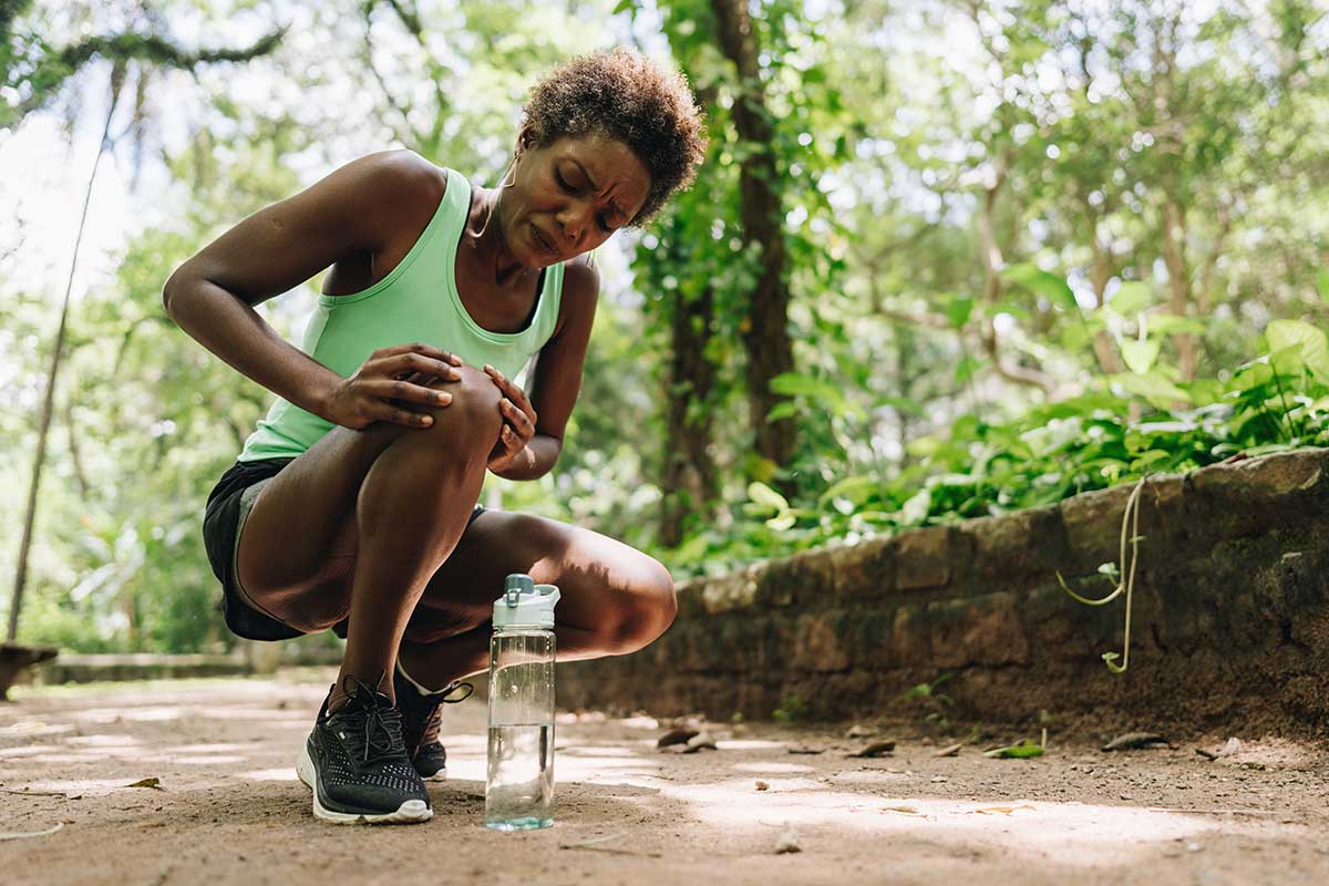 A woman on a wooded trail crouches and holds her knee in apparent pain. She is wearing shorts and a green top and has placed her water bottle on the ground next to her.